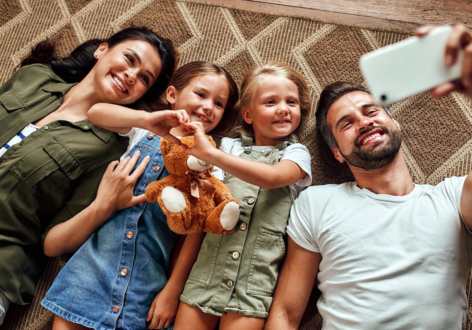 Dad, mom and two cute daughters lie on the carpet on the floor in the living room and take a selfie on a smartphone. Happy family have fun together.